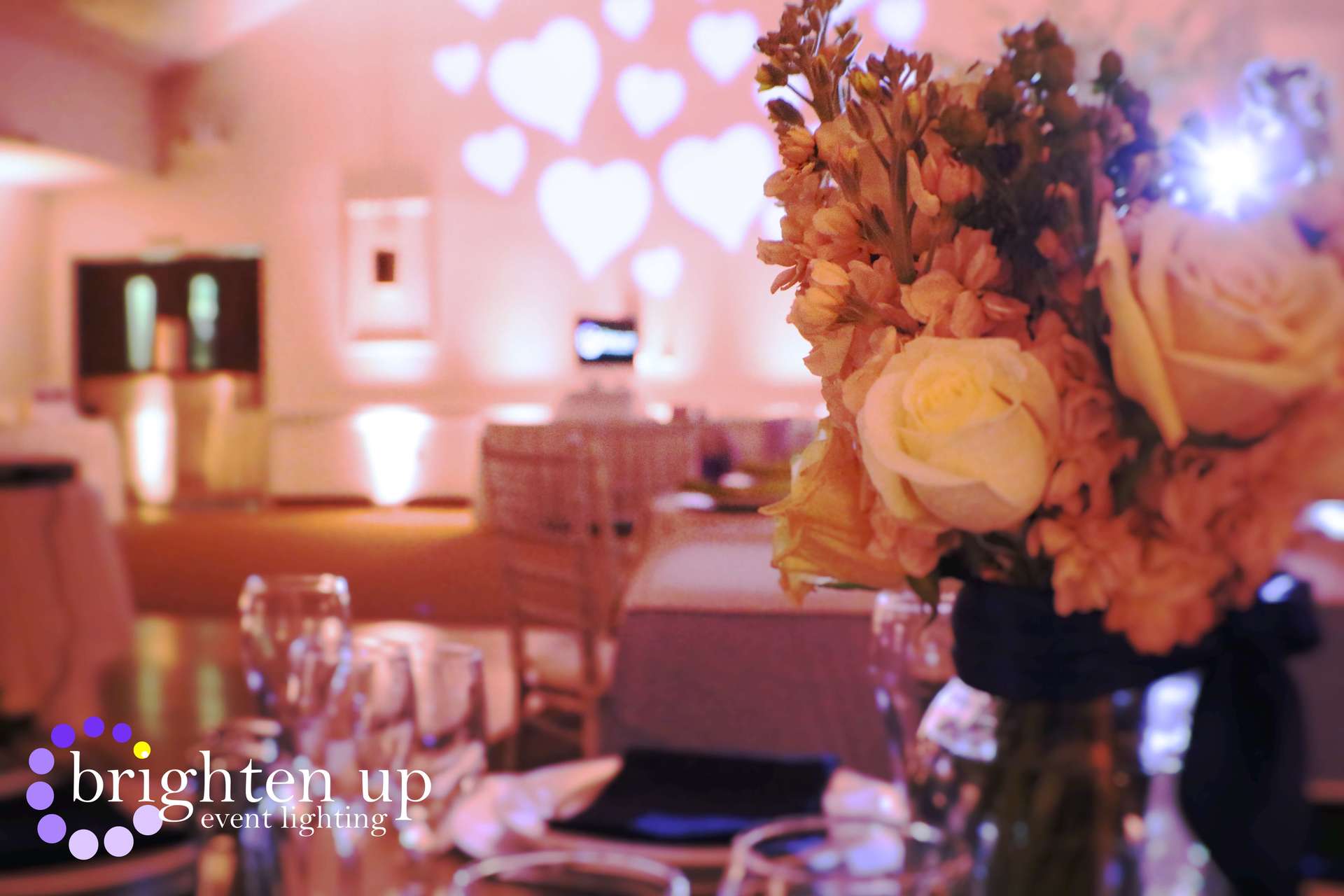 A vase of flowers sits on a table with hearts projected on the wall behind it.