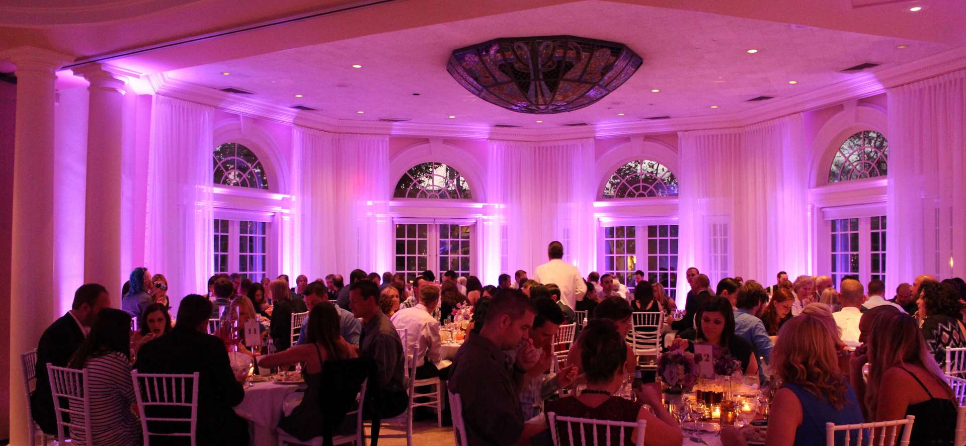A large group of people are sitting at tables in a large room with purple lights.