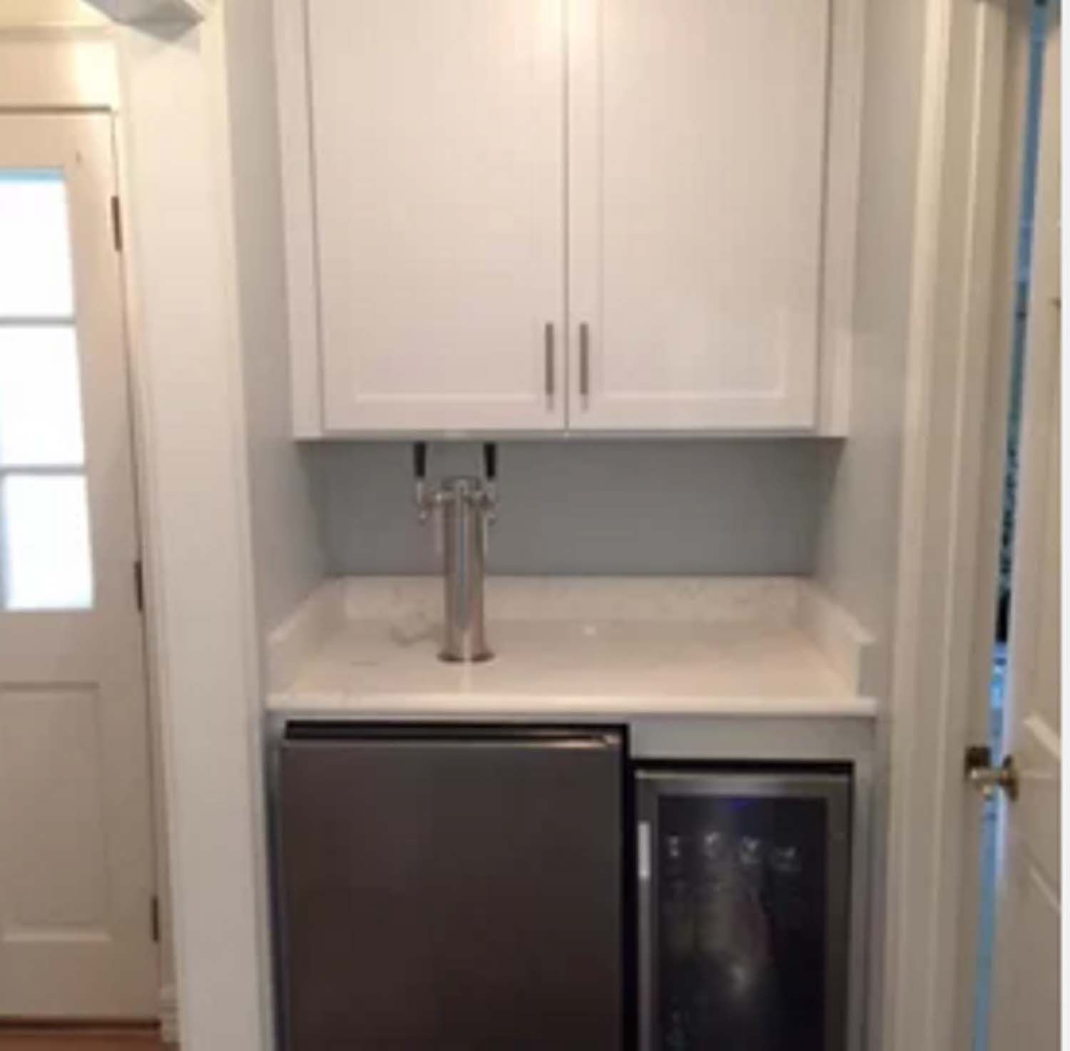 White cabinet with beer tap, built over a small fridge and wine cooler. Doorway on the left.