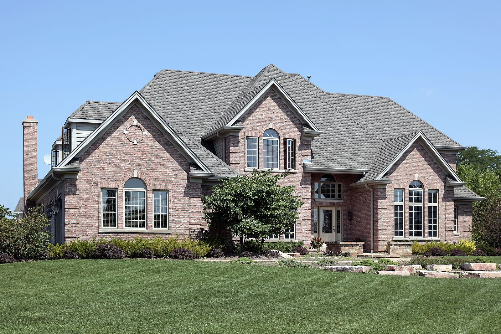 Brick house with a gray roof, multiple windows, and a well-manicured lawn under a blue sky.
