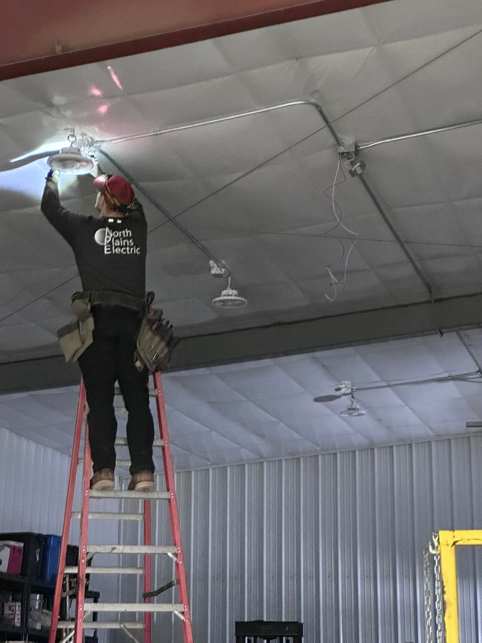 Electrician on ladder, installing ceiling light in metal building.