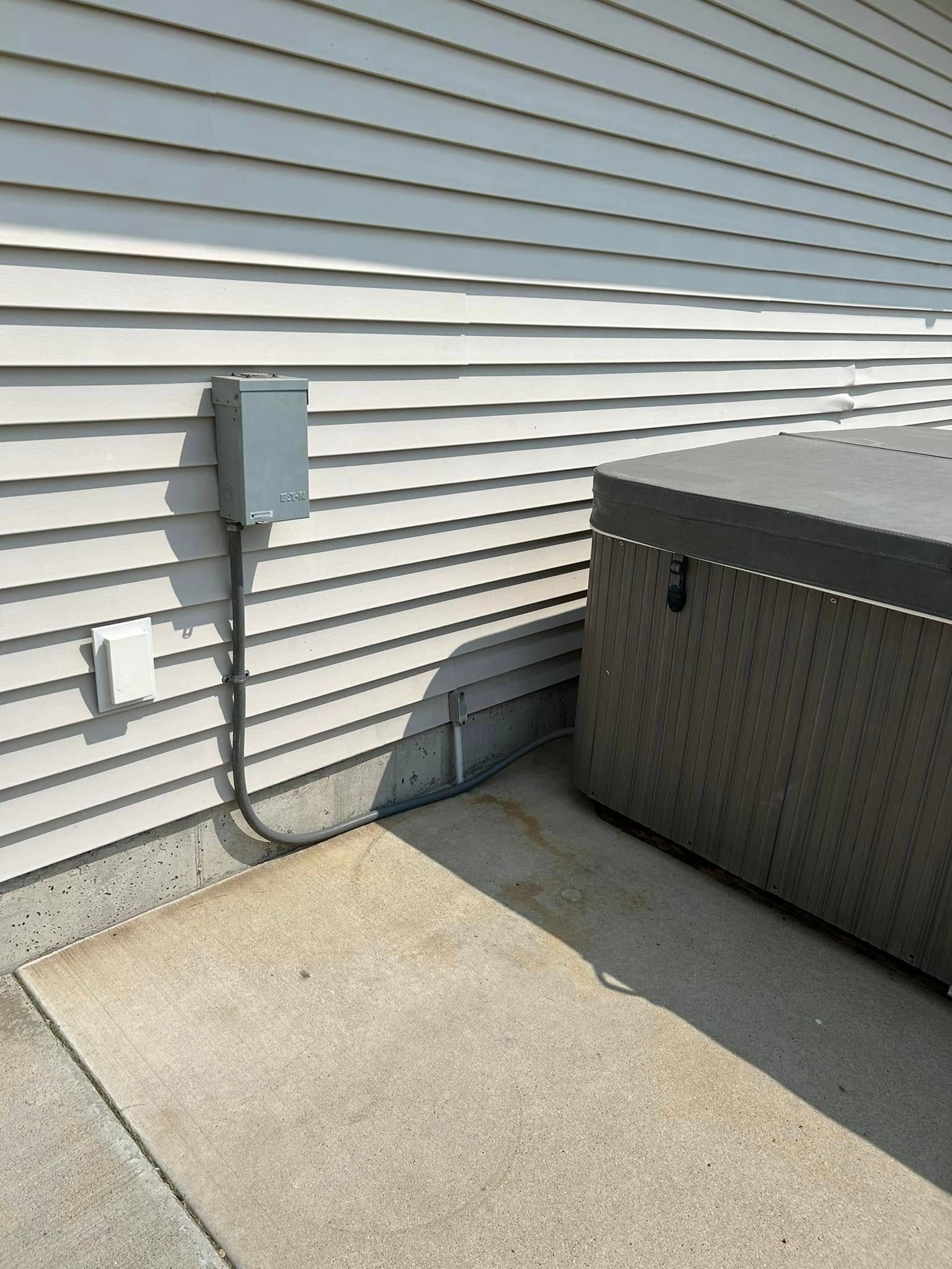 Gray electrical box and outlet on a house exterior next to a hot tub on a concrete pad.