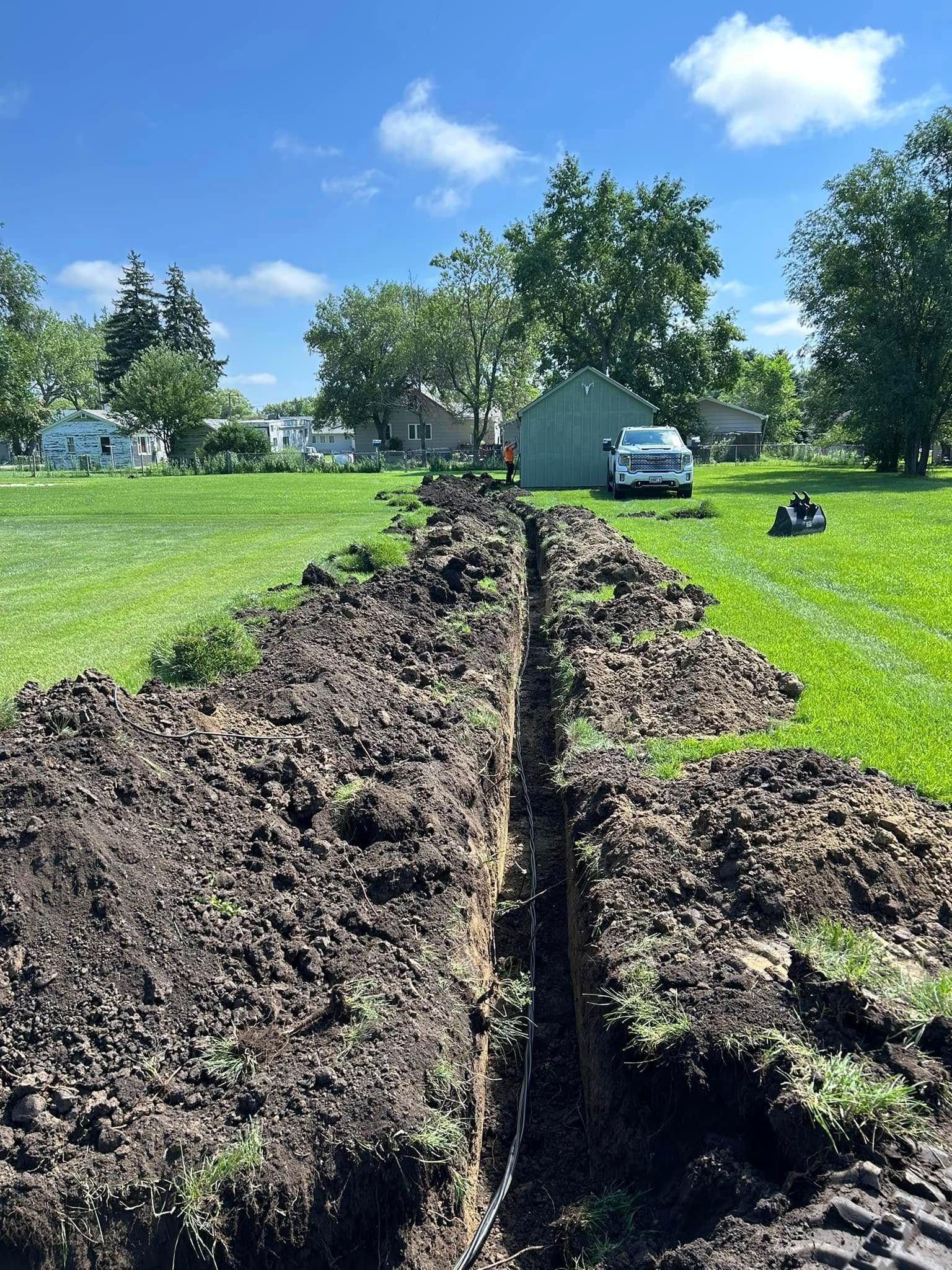 Trench dug in a grassy field, leading to a shed and work truck under a blue sky.