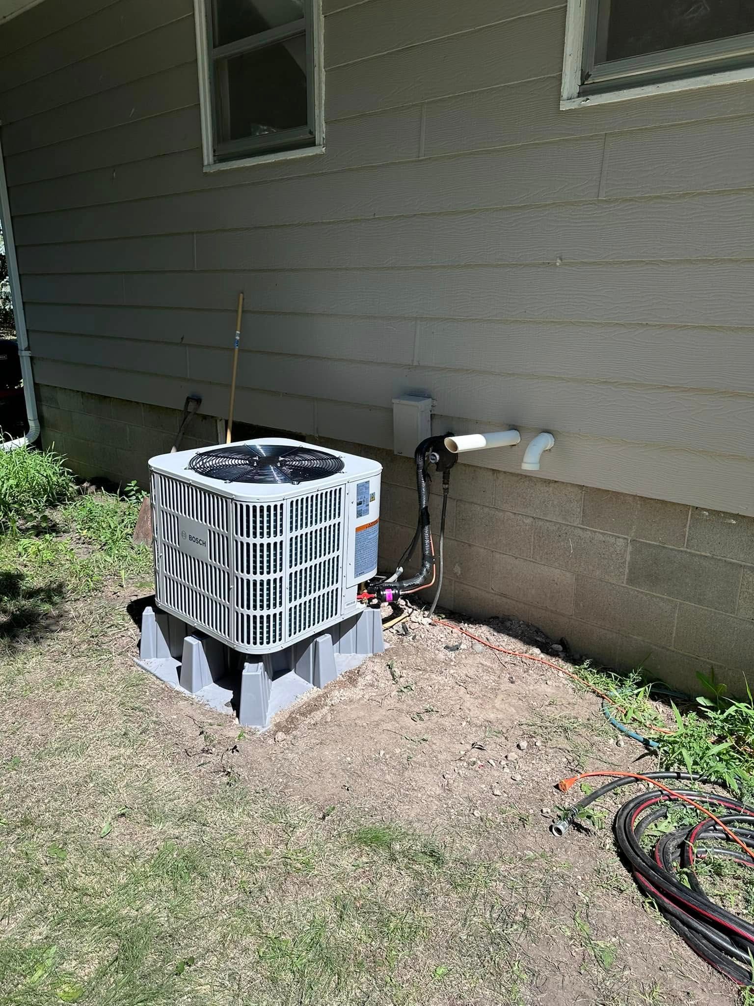 Central air conditioning unit outside a light-colored house. Unit sits on blocks. Green grass around it.