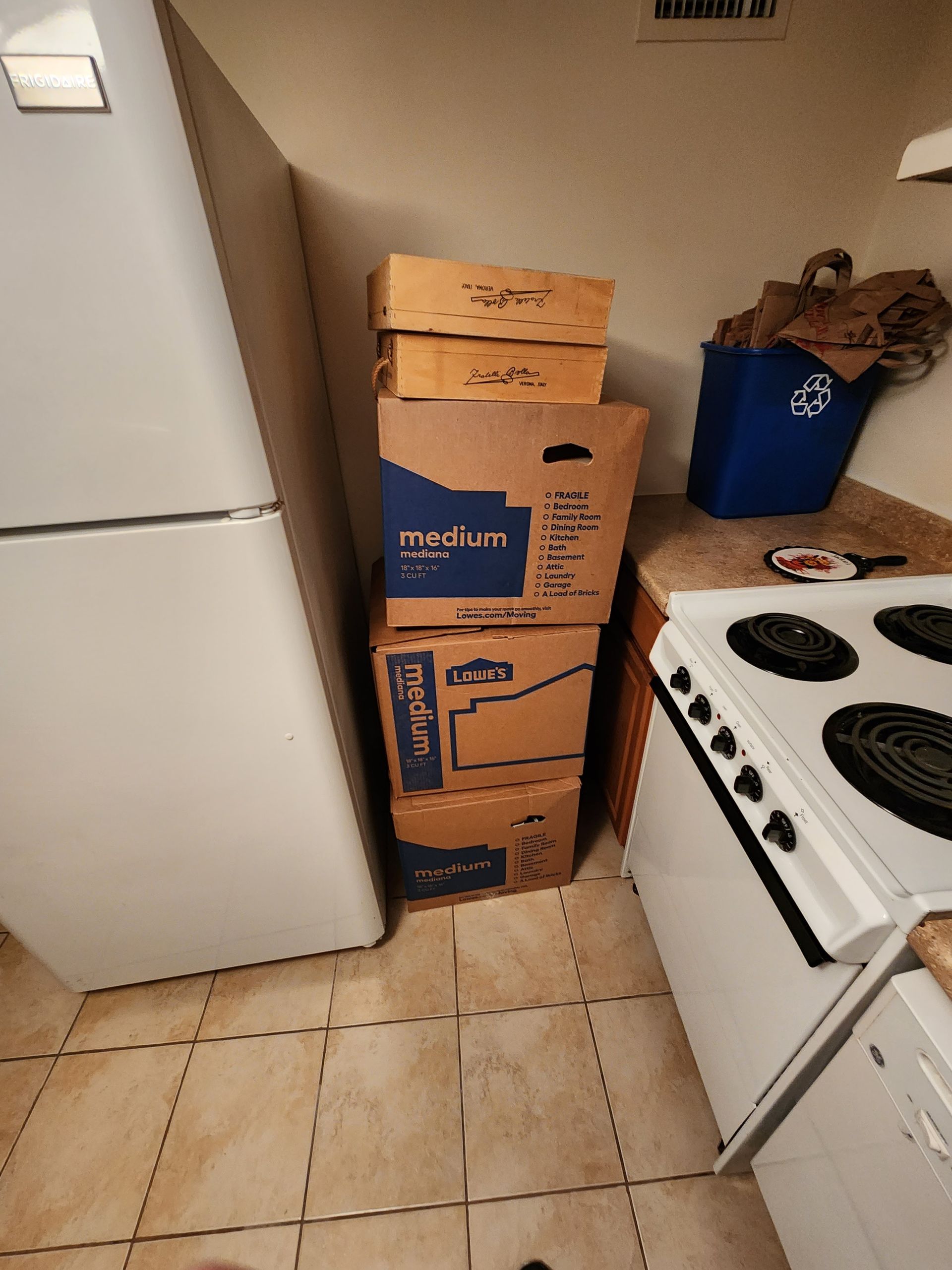 A stack of cardboard boxes in a kitchen next to a stove and refrigerator.