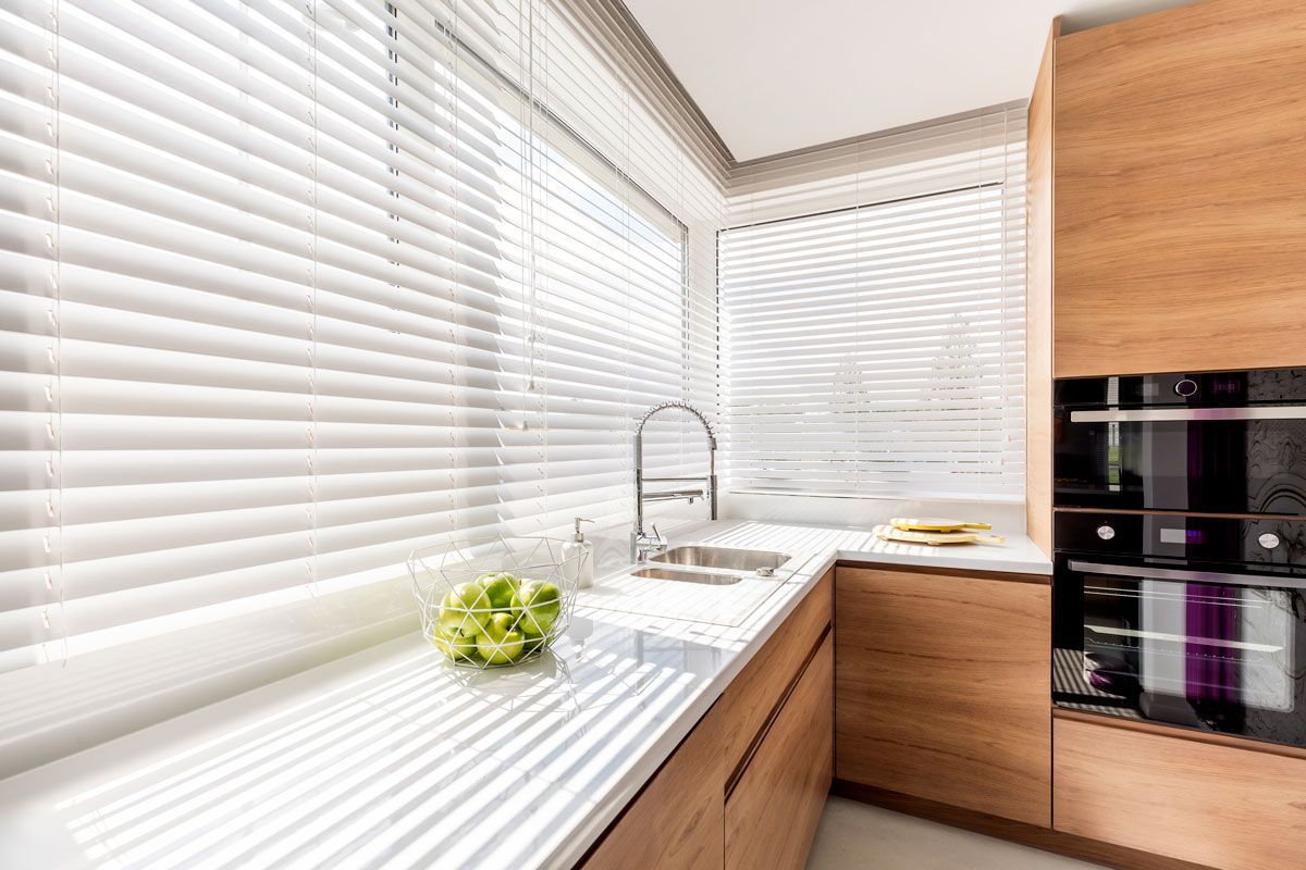 A bright kitchen interior with white window blinds, and wooden cabinets with white countertop.