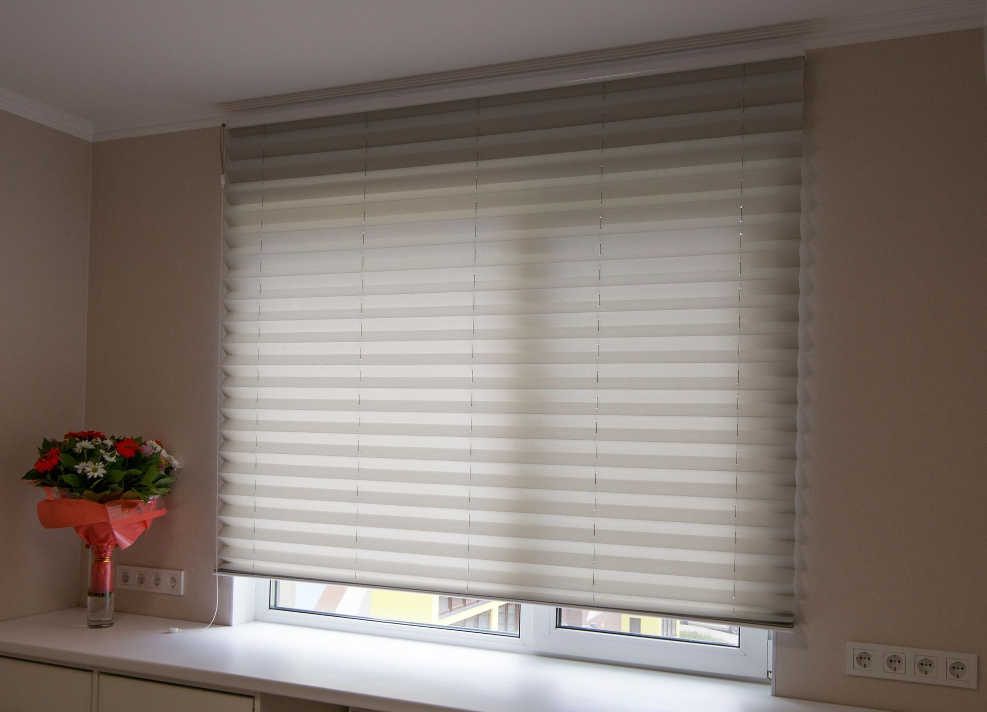 Closed layered window blinds above a sill with flowers and power outlets inside a room.