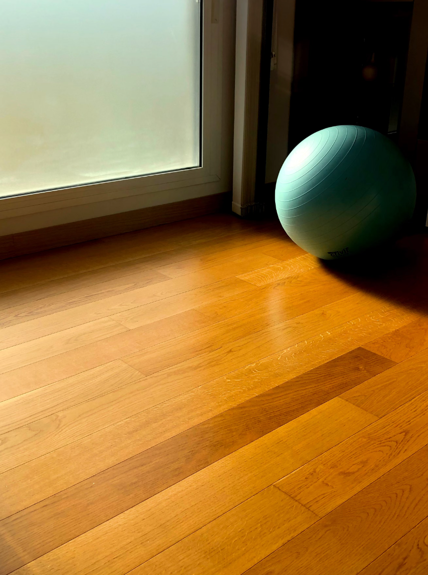 A green exercise ball is sitting on a wooden floor next to a window.