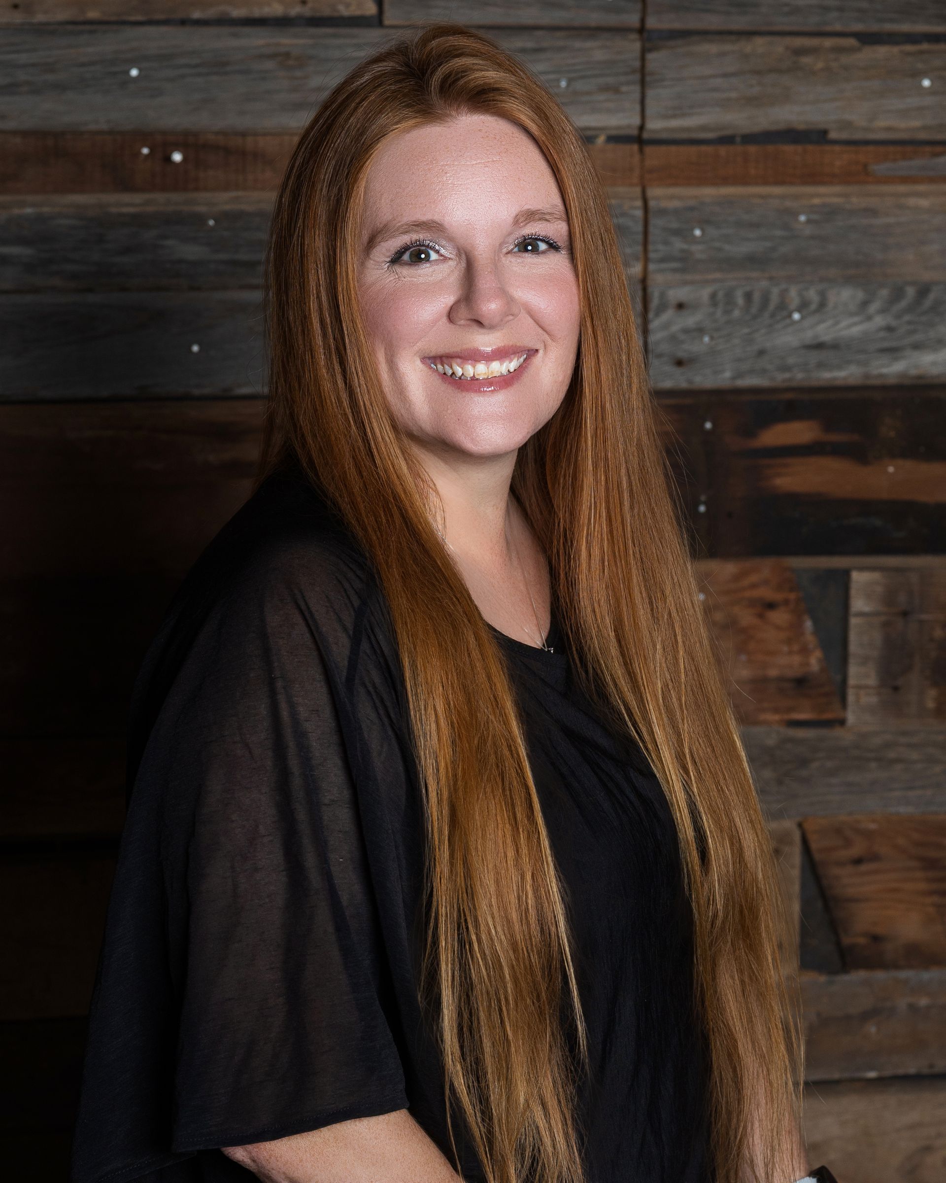 A woman with long red hair is smiling in front of a rusty metal wall.