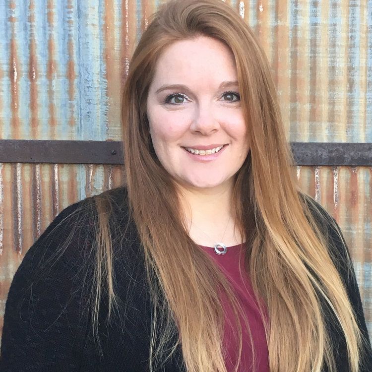 A woman with long red hair is smiling in front of a rusty metal wall.