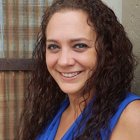 A woman with curly hair is wearing a blue shirt and smiling.