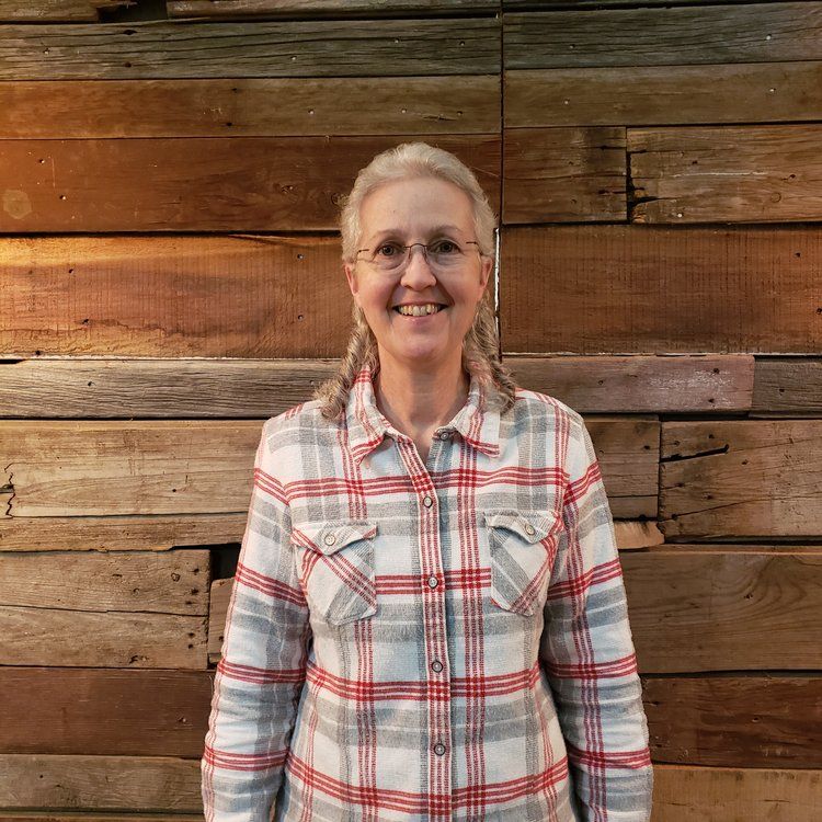 A woman wearing glasses and a plaid shirt is standing in front of a wooden wall.