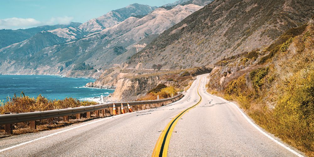 A road going through a valley with mountains and the ocean in the background.