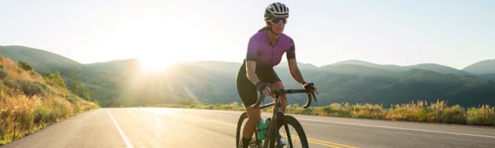A cyclist in a purple jersey rides a road bike along an open highway at sunrise with mountain ranges in the distance.