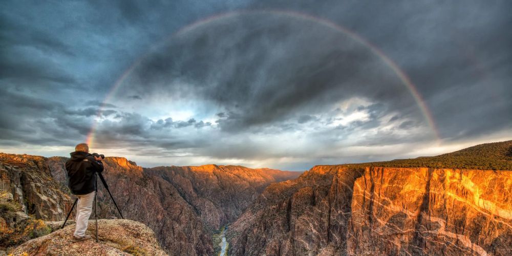 A man is taking a picture of a rainbow over a canyon.