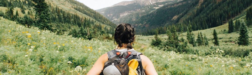 A woman with a backpack is walking through a grassy field in the mountains.