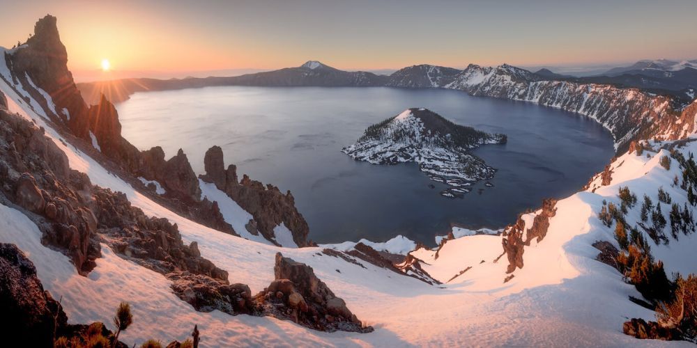 The sun is setting over a lake surrounded by snow covered mountains.