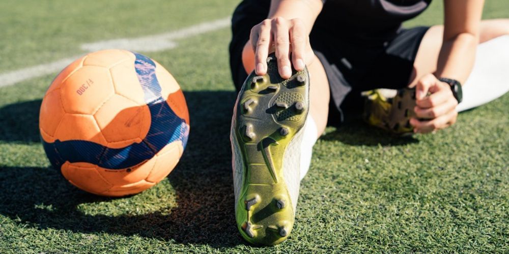 A person is stretching their legs on a soccer field next to a soccer ball.