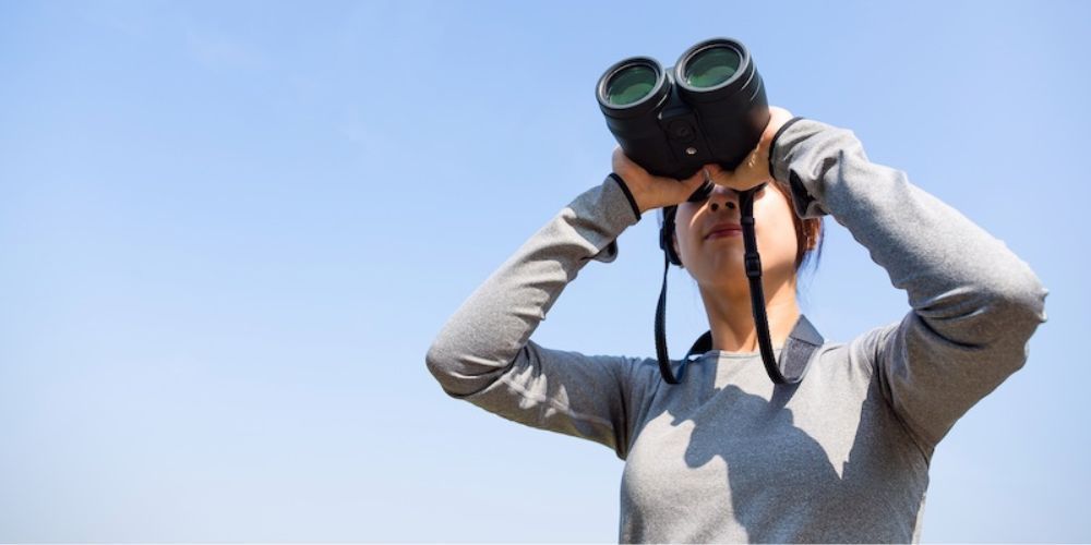 A woman is looking through binoculars against a blue sky.