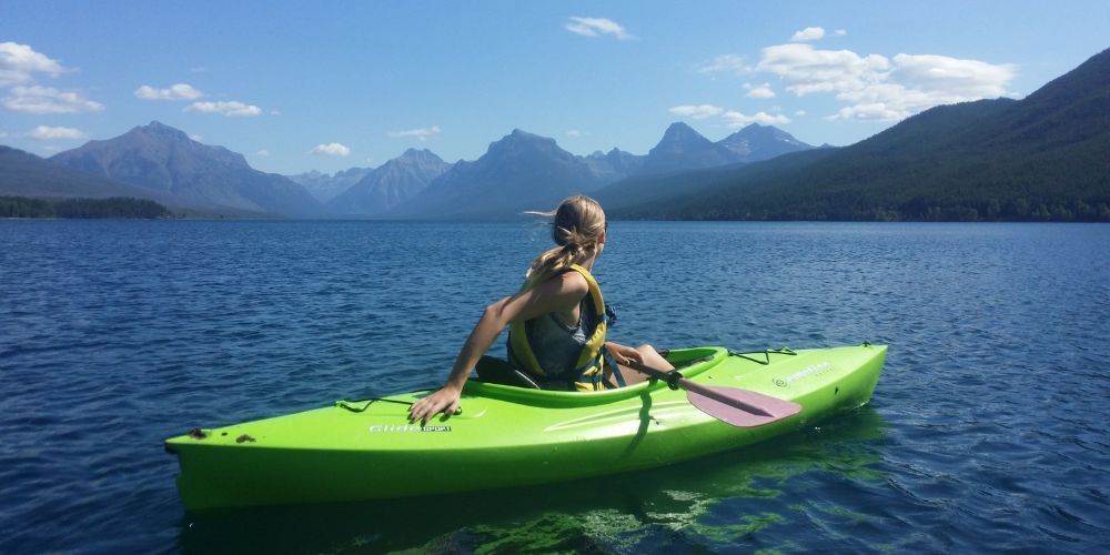 A woman is sitting in a green kayak on a lake with mountains in the background.