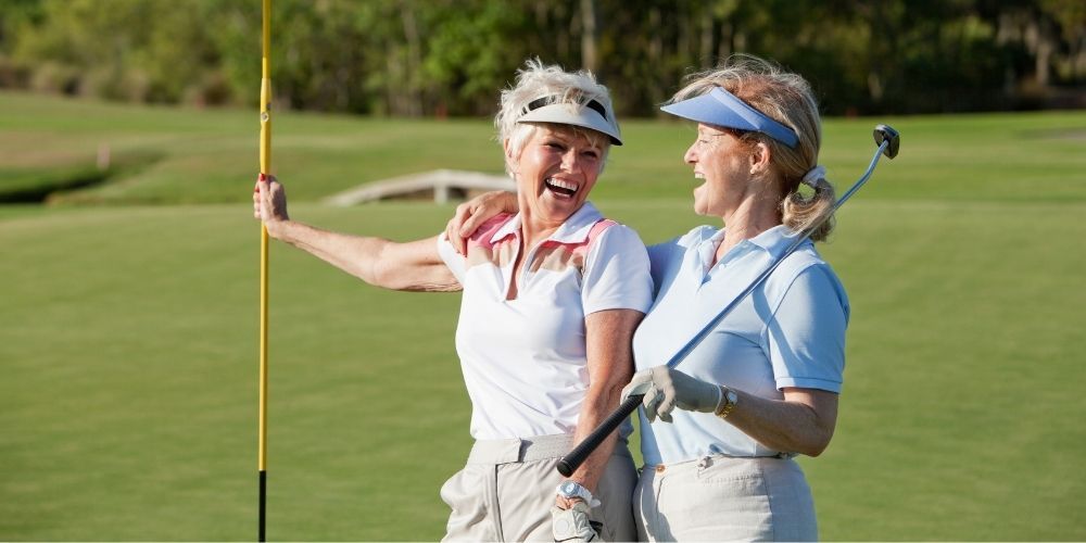 Two older women laughing on a golf course. One holds a golf flag, the other holds a golf club. They are wearing visors and light-colored clothing.