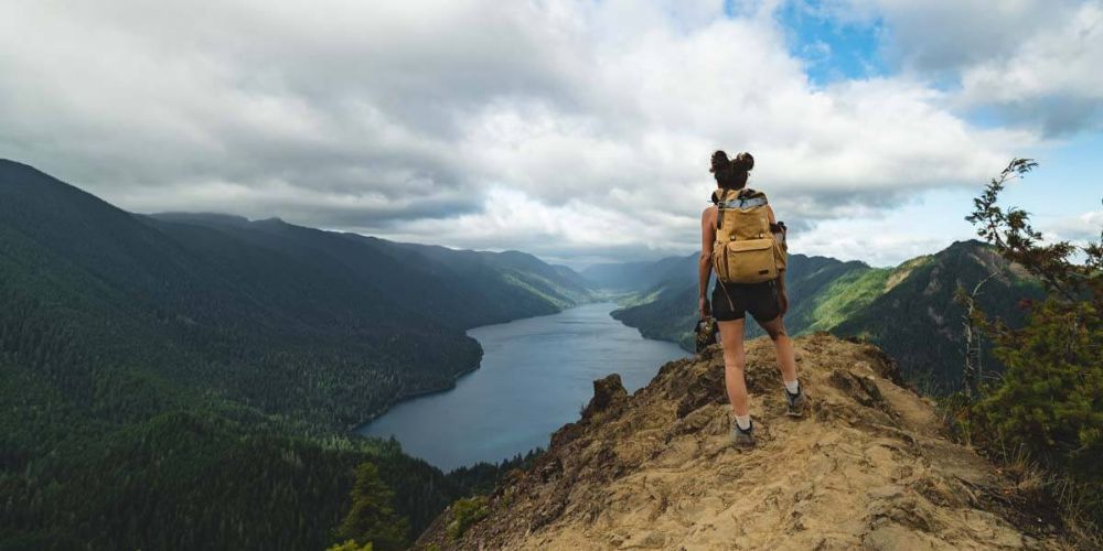 A woman with a backpack is standing on top of a mountain overlooking a lake.