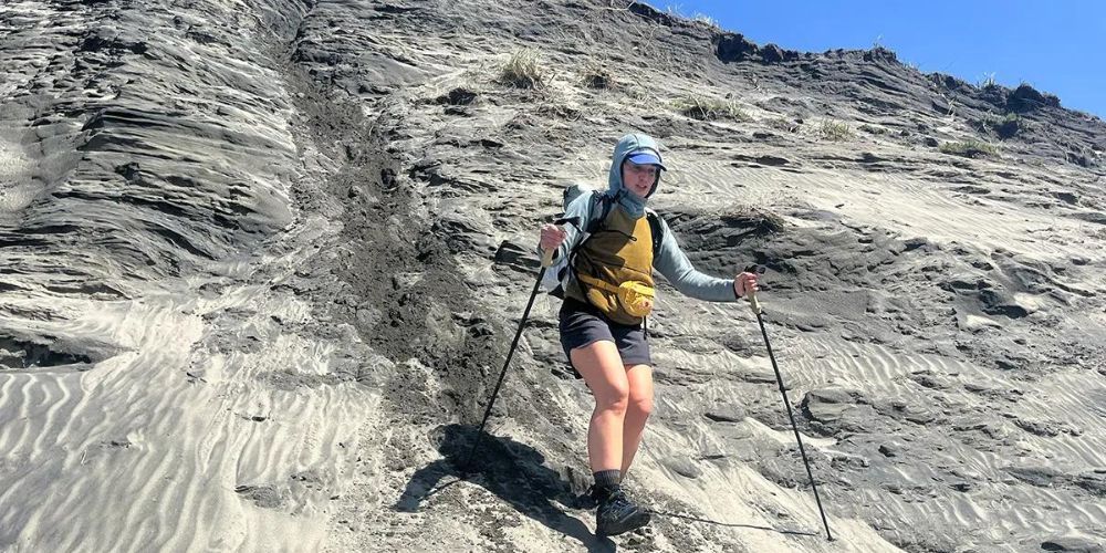 A woman is hiking up a sandy hill with hiking poles.