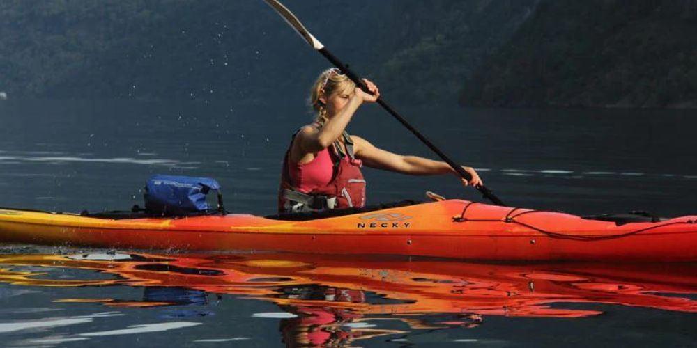 A woman is paddling a red and yellow kayak on a lake.