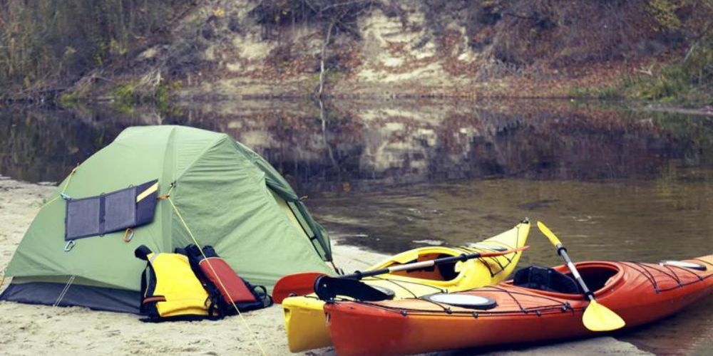 A tent and two kayaks are on the beach next to a river.