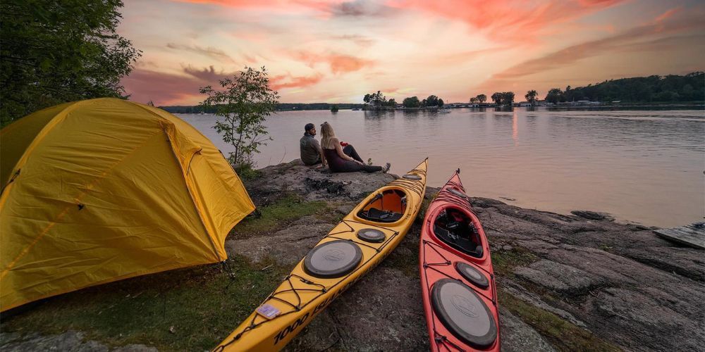 Two kayaks are sitting on the shore of a lake next to a tent.