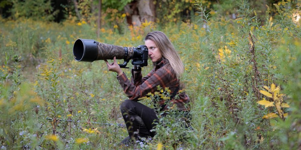 A woman is kneeling down in a field holding a camera.