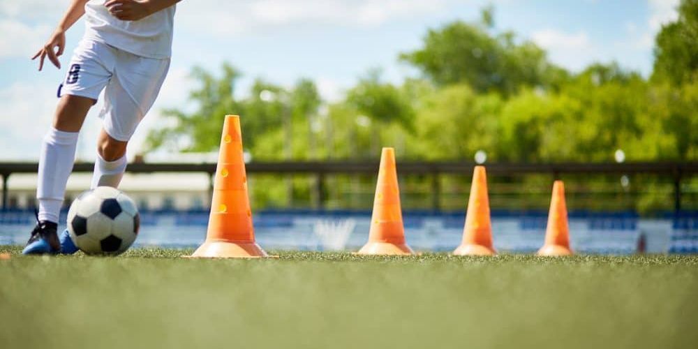 A soccer player is kicking a soccer ball between orange cones on a field.