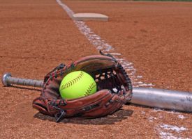 A softball in a glove and a bat on a softball field.