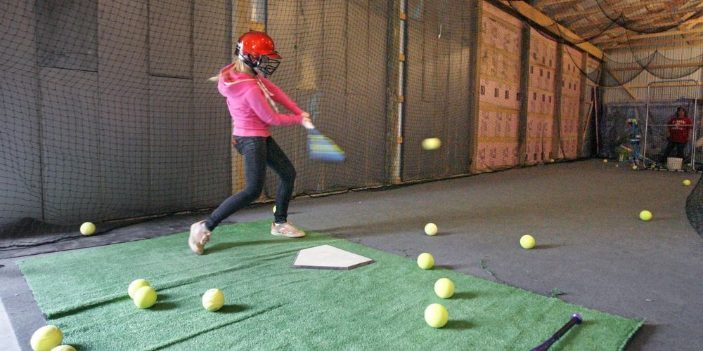 A young girl is swinging a bat at a baseball in a cage.