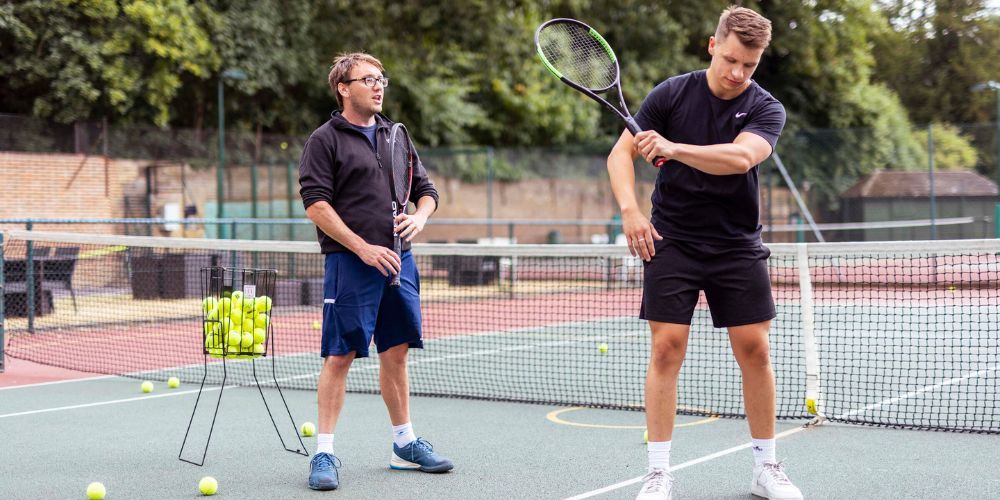 A man is teaching another man how to play tennis on a tennis court.