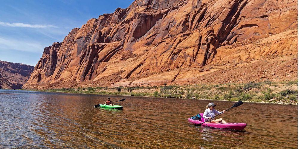 Two people are kayaking down a river in front of a rocky cliff.