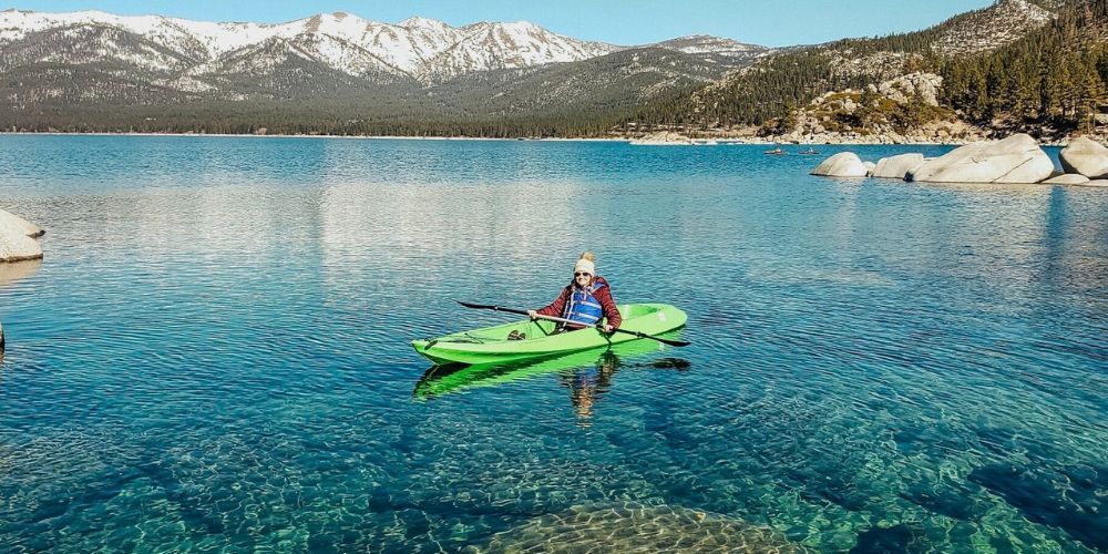 A person is riding a kayak on a lake with mountains in the background.