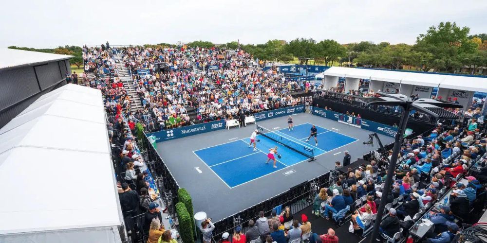 A crowd of people are watching a pickleball match on a tennis court.