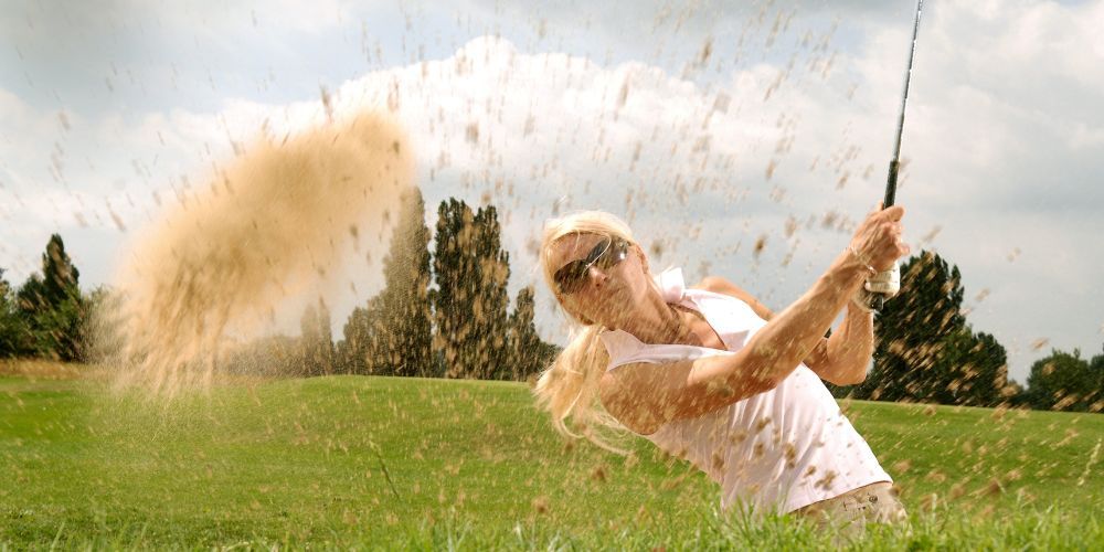 A blonde woman in sunglasses swings a golf club from a sand trap, kicking up sand and grass.