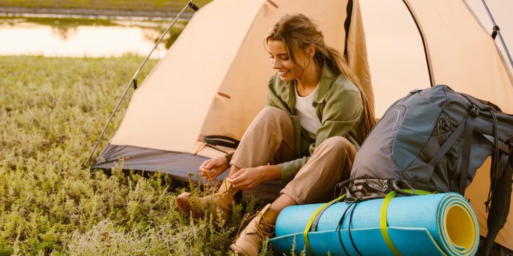 Woman smiles while tying her boots, sitting outside a tent in a grassy field. Camping gear nearby.