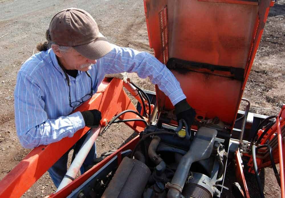 Man in Hat and Gloves Working on Tractor Engine Outdoors — Paul’s Tyres & Farm Maintenance in Dubbo, NSW