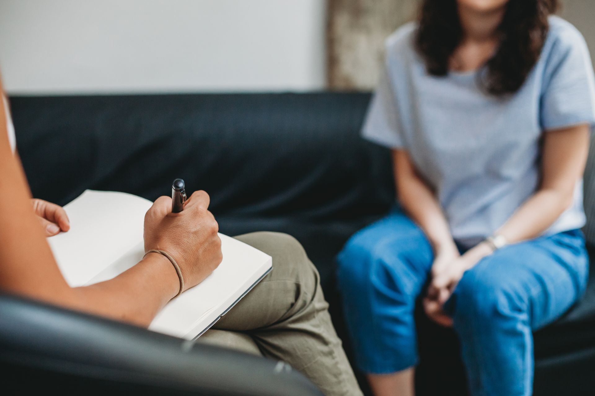 A woman is sitting on a couch talking to a man who is writing in a notebook.