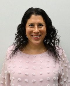 A woman in a purple floral shirt is smiling in front of a bush.