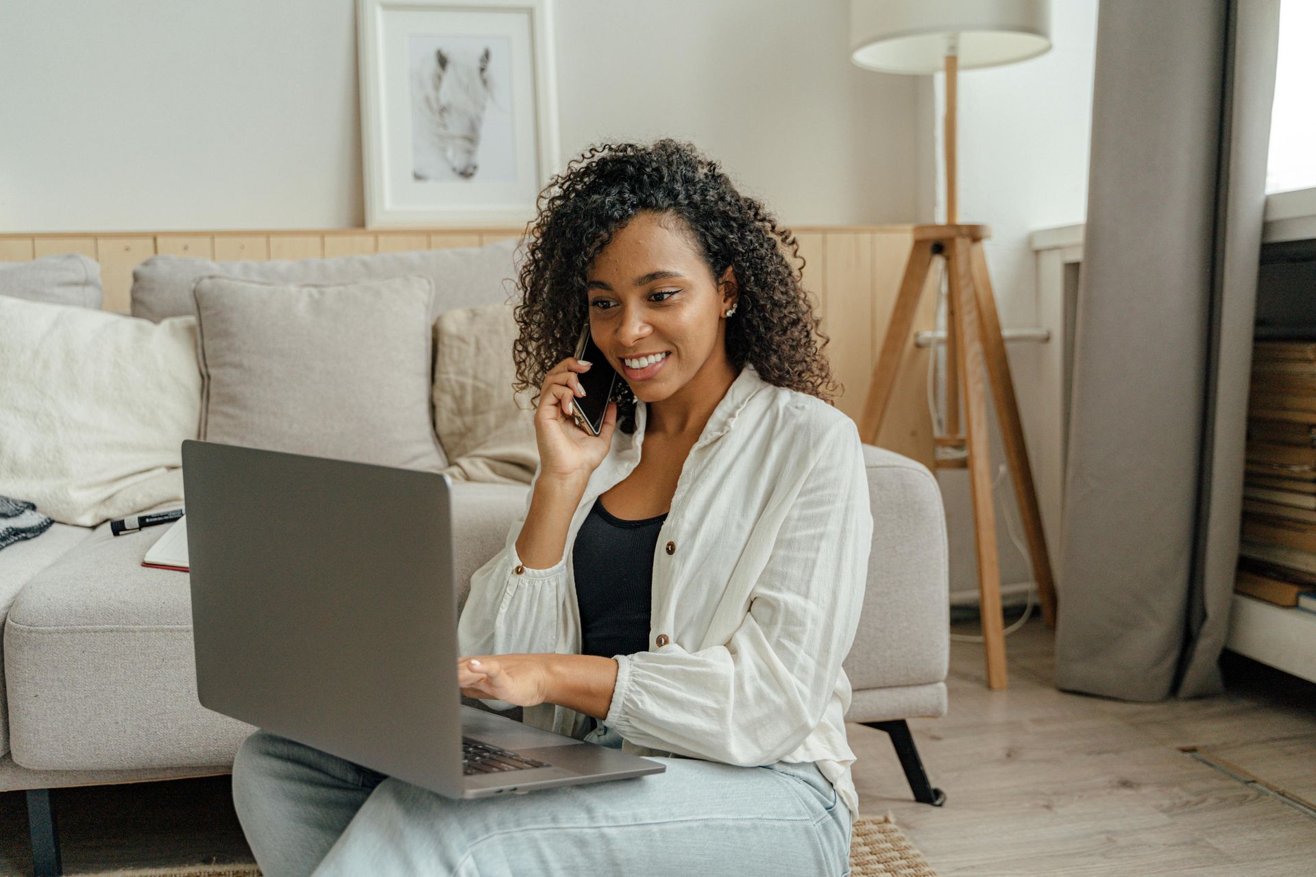 A woman is sitting on the floor using a laptop and talking on a cell phone.