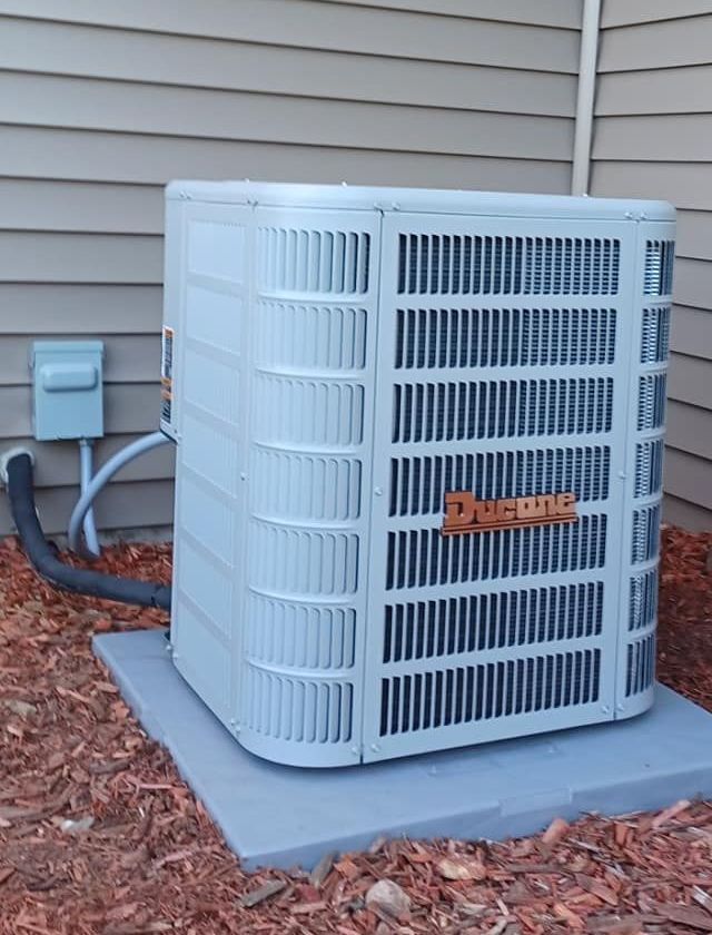 An outdoor air conditioning unit, light blue, on a gray concrete pad, next to a tan siding wall. The unit has the Ducone brand name.