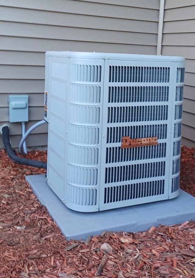 An outdoor air conditioning unit, light gray with vertical slats, sits on a concrete pad against a beige house with brown mulch.
