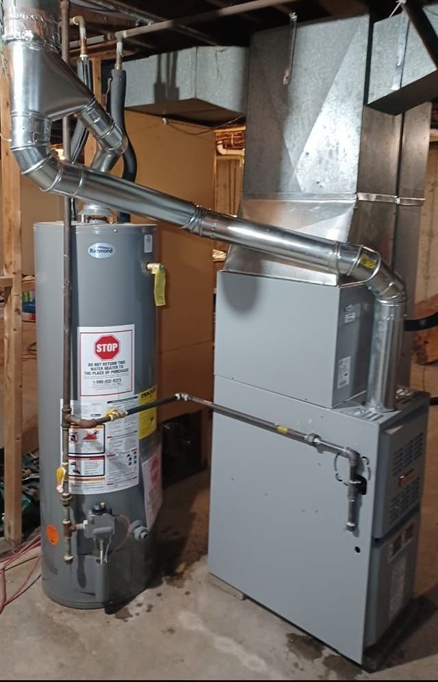 A water heater and furnace sit side-by-side in a basement. Silver ducts connect the appliances. The water heater is gray, and the furnace is light gray.