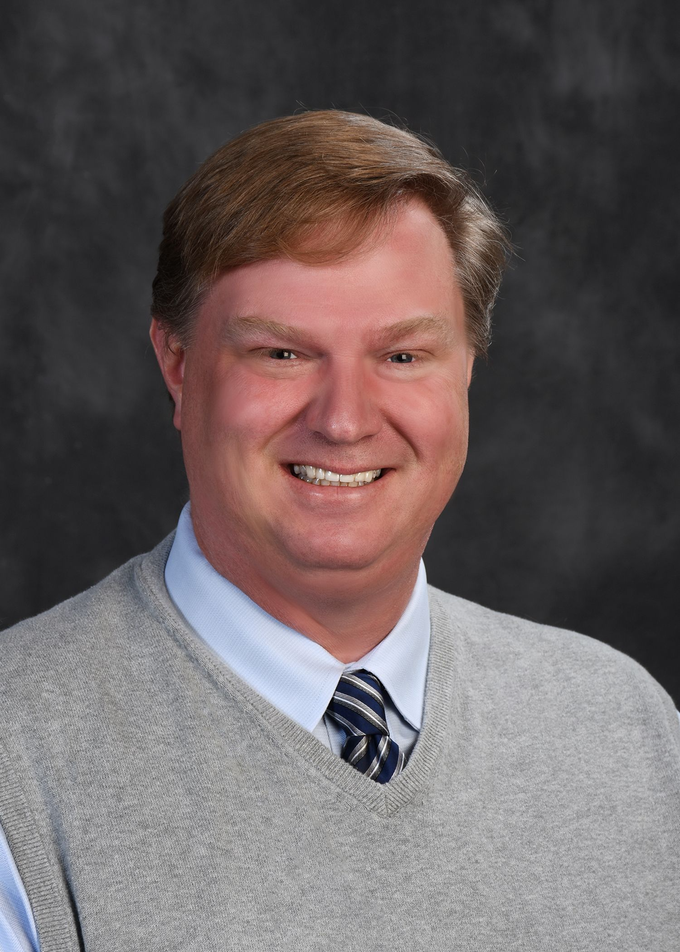 Man in a light gray sweater smiling, blue collared shirt and tie against a gray background.