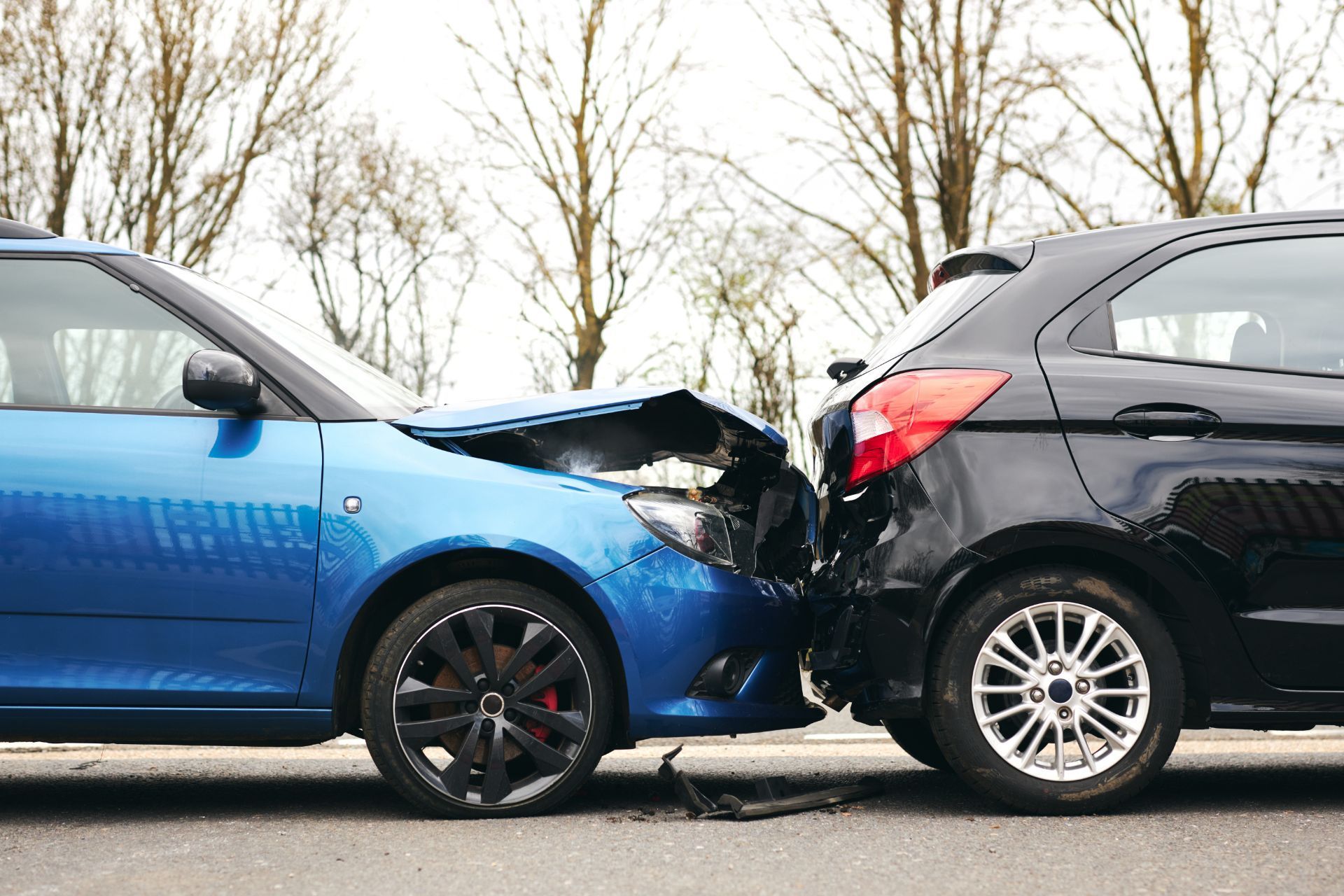 Two cars, blue and black, crashed on a road. The blue car's front is damaged, and the black car's rear is damaged.