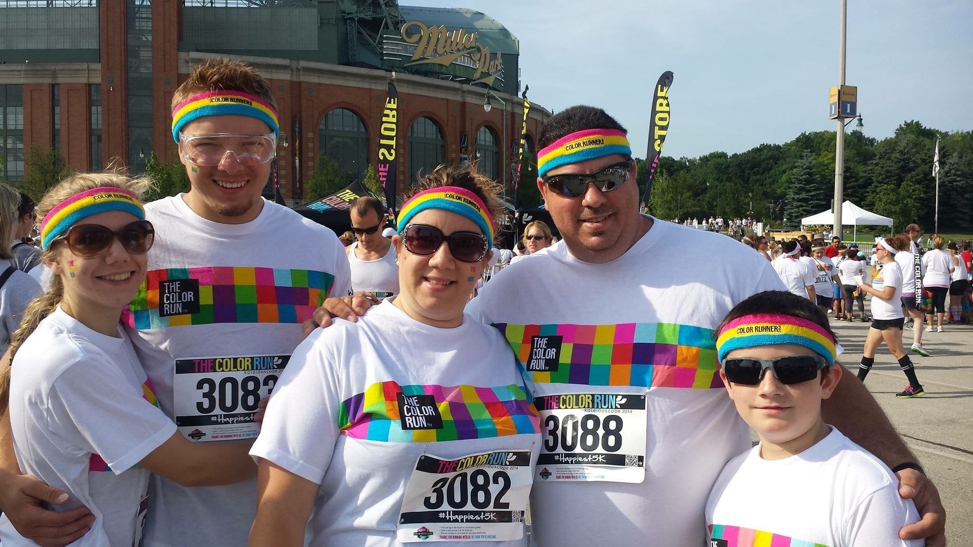 Five people in white t-shirts and rainbow headbands pose together. They are in front of a stadium during a color run.